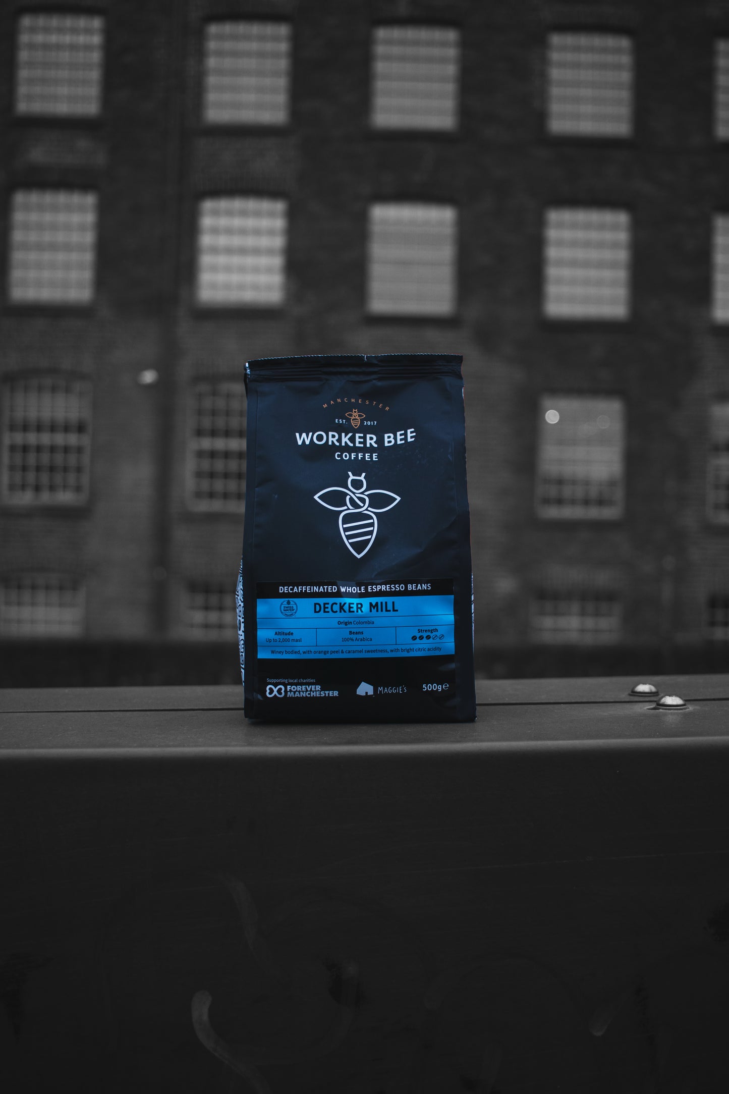 A bag of Worker Bee Tea & Coffee’s Decker Mill Decaf Espresso Beans – Swiss Water 500g sits outdoors, its dark package with blue and white text and a bee logo highlighting its chemical-free Swiss Water decaf process, with a brick building in the background.