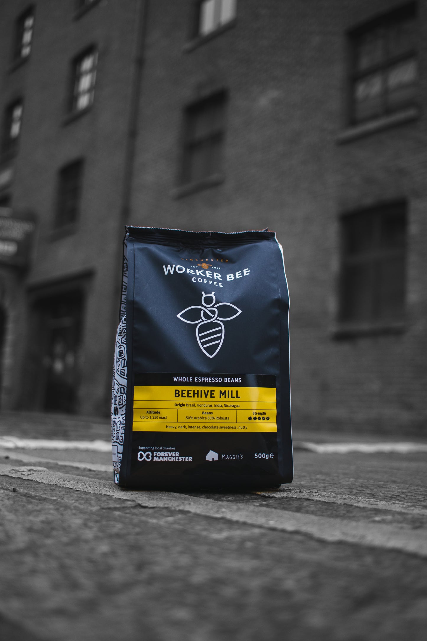A bag of Worker Bee Tea & Coffee Beehive Mill Espresso Beans 500g sits on a street with blurred brick buildings behind it. The black bag has a yellow label, a bee logo, and contains a 50/50 blend of Arabica and Robusta beans.